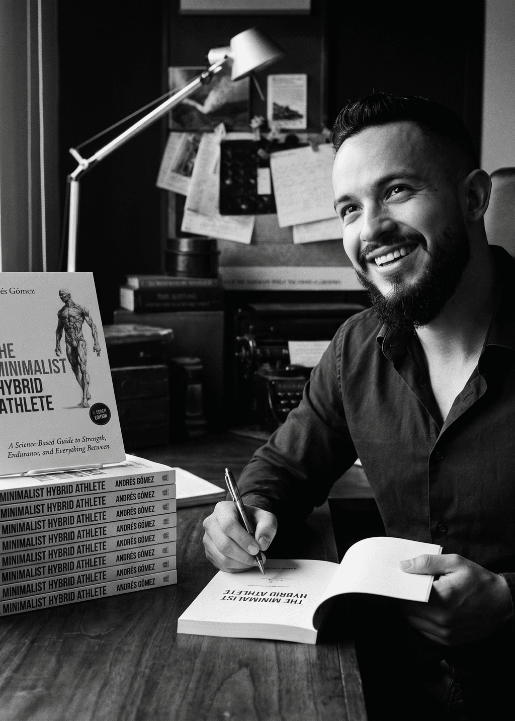 Andrés Gómez smiling as he signs copies of his book The Minimalist Hybrid Athlete at a desk stacked with copies — monochrome editorial portrait